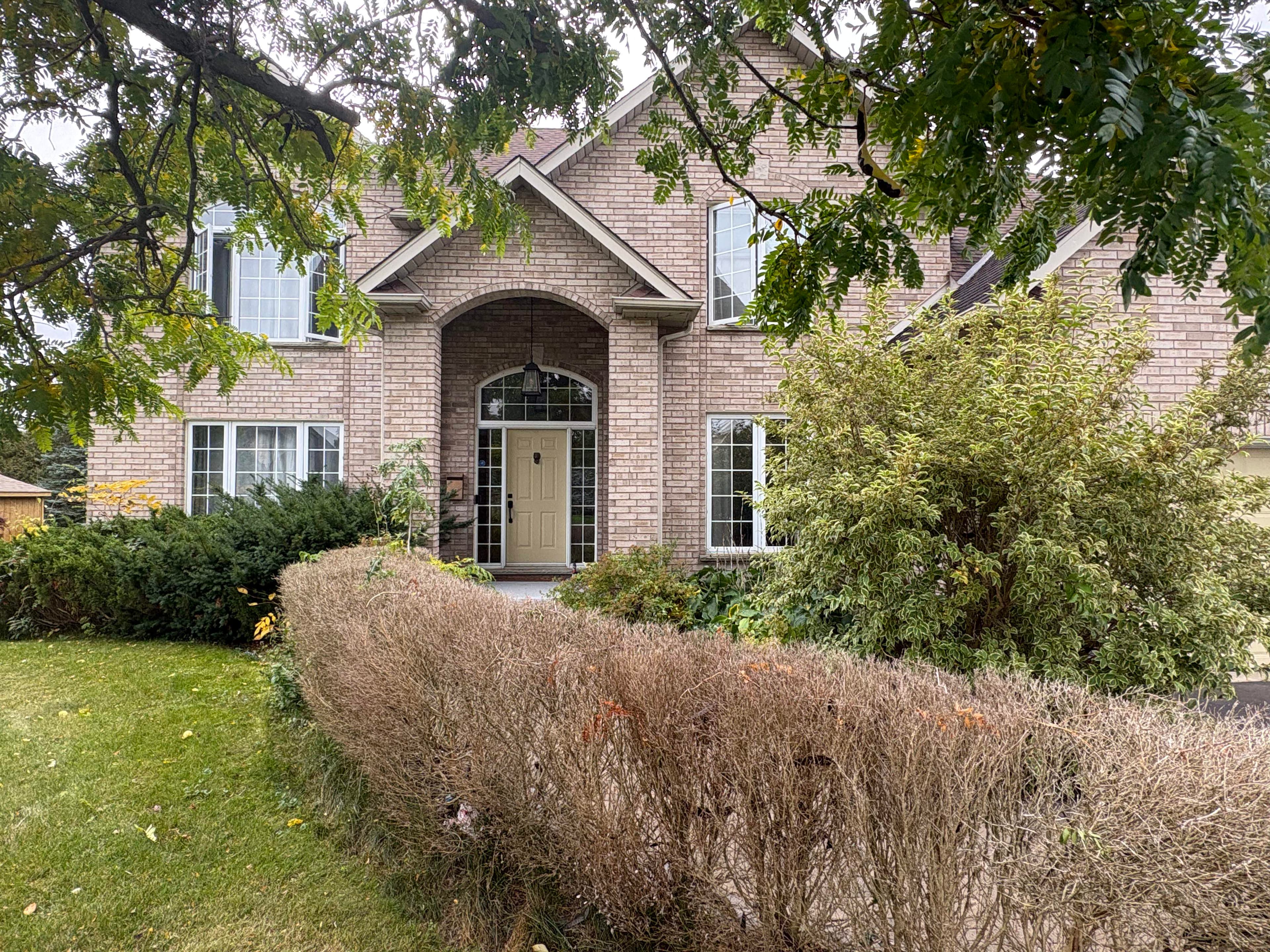 Two-story light brick house with arched entryway and white windows behind a dry hedge.