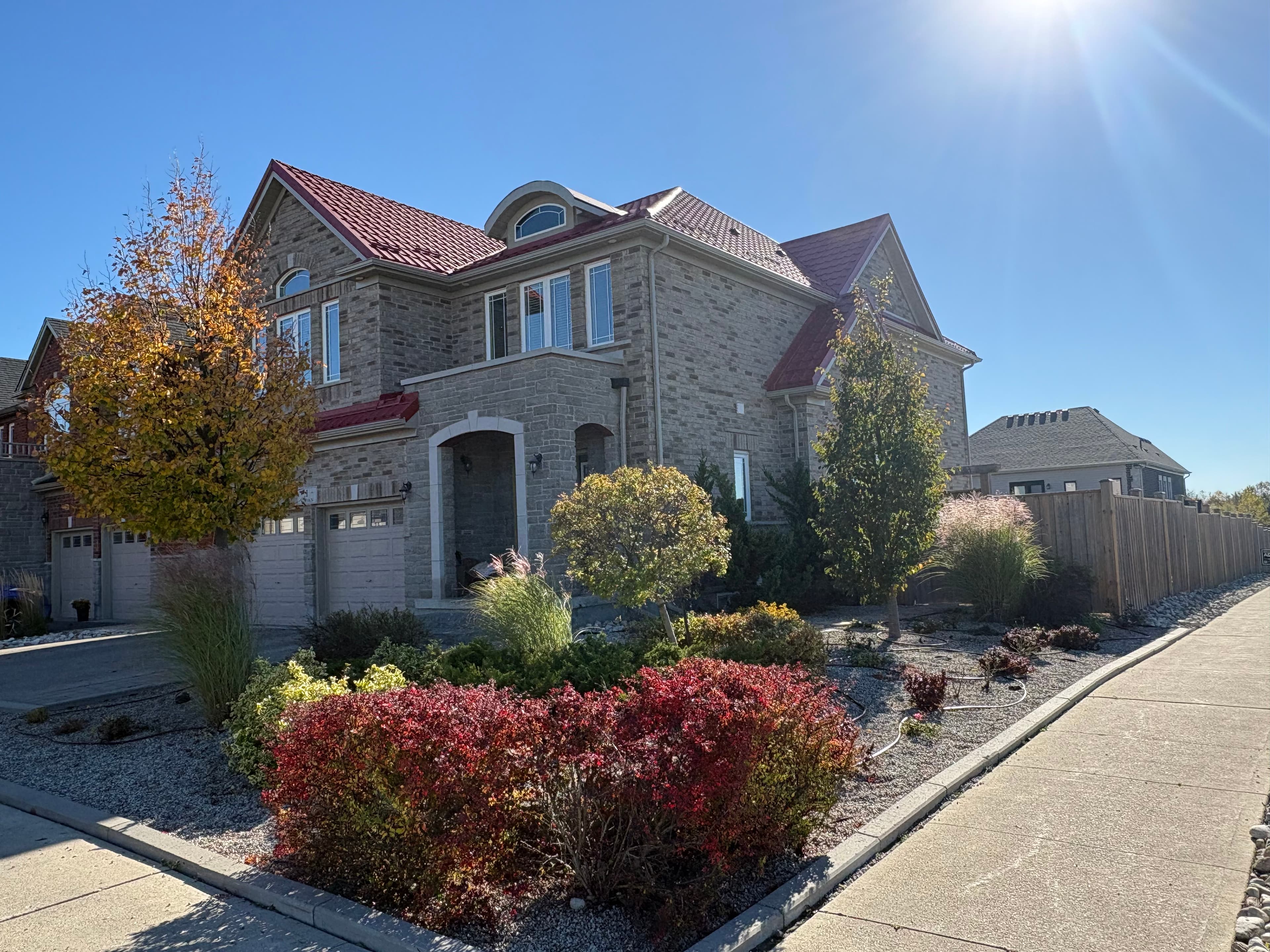 Large stone house with a red metal roof and colorful landscaping under a bright sun.